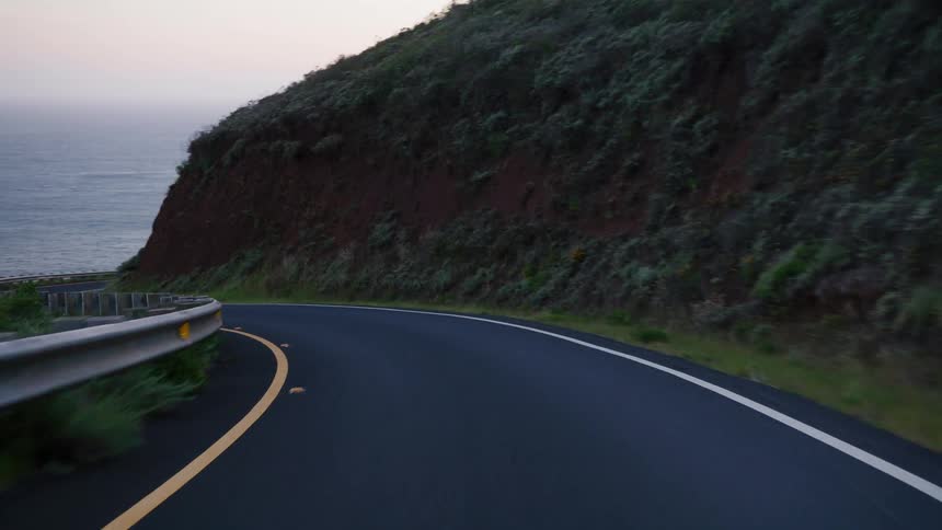 A point-of-view shot driving along a winding coastal road next to the ocean at dusk
