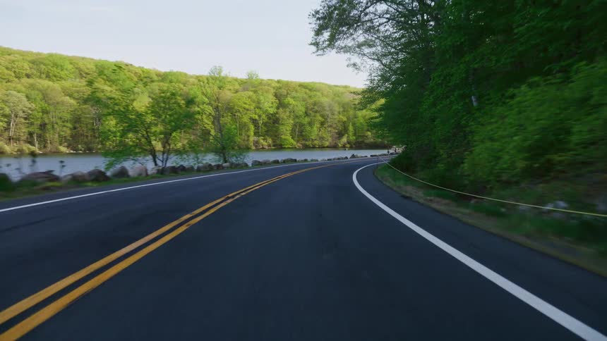 A scenic point-of-view drive on a winding country road alongside a calm lake surrounded by a lush green forest
