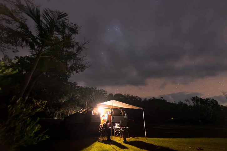A stunning time-lapse transitions from a colorful sunset to a brilliant night sky full of stars and the Milky Way over a campervan at a campsite