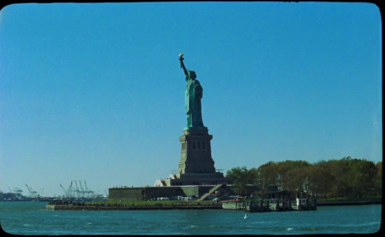 A grainy, vintage 16mm film captures the Statue of Liberty standing proudly in New York Harbor on a clear, sunny day