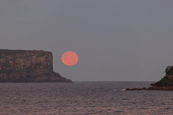 A stunning day-to-night timelapse captures a vibrant supermoon rising over the ocean between coastal cliffs as boats sail by