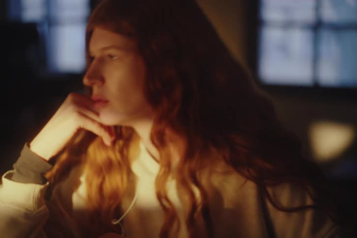A contemplative young woman with long red hair listens to music with earphones while sitting in a dimly lit room
