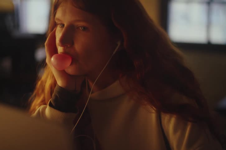Slow motion close-up of a young woman with red hair blowing a bubble gum bubble that pops on her face