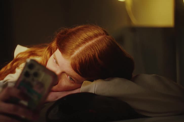 A melancholy young woman with red hair lies in bed in her dimly lit room at night, looking at her phone