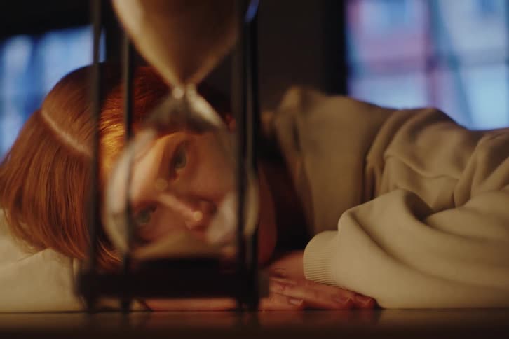 A close-up shot of an hourglass with a pensive red-haired woman coming into focus in the background, contemplating the passage of time