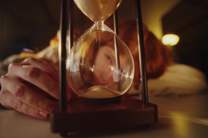 A dramatic close-up of an hourglass with flowing sand, reflecting a still person lying in a dimly lit room as time runs out