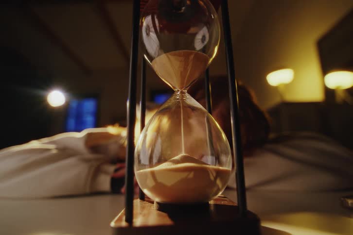 An hourglass marks the passage of time with falling sand as a person watches intently in a dimly lit, atmospheric room