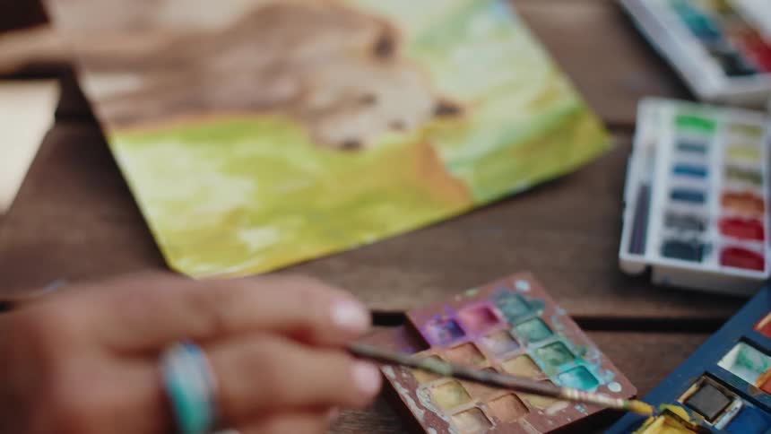An artist's hand dips a paintbrush into a vibrant watercolor palette and adds color to a painting in a slow-motion close-up