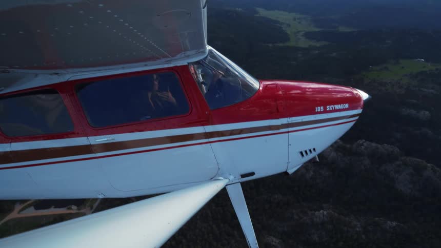 Cinematic aerial shot of a red and white Cessna 185 Skywagon with a pilot and passenger flying over a scenic mountain range