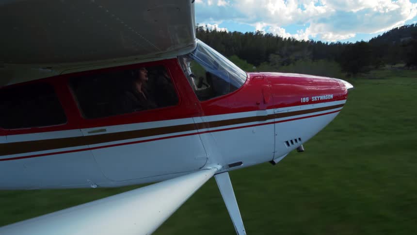 View from the wing of a small red and white plane as a female pilot expertly lands on a grassy airstrip in a remote mountain valley