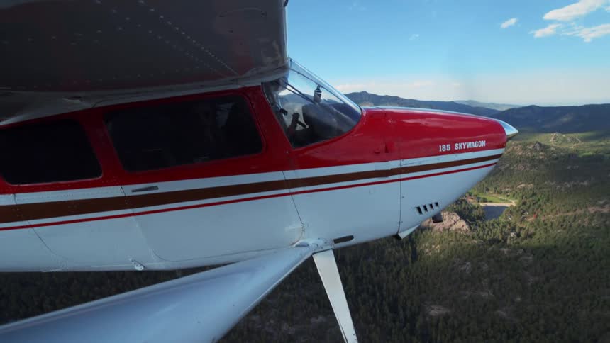 A red and white 185 Skywagon single-engine airplane soars over a vast mountain range covered in lush green forests on a clear day