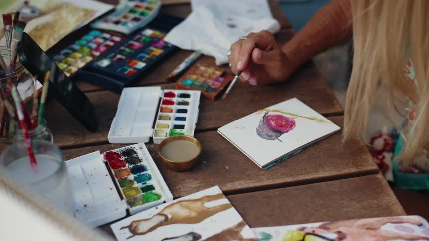 Close-up of a female artist's hands painting a watercolor still life on a rustic wooden table surrounded by her art supplies