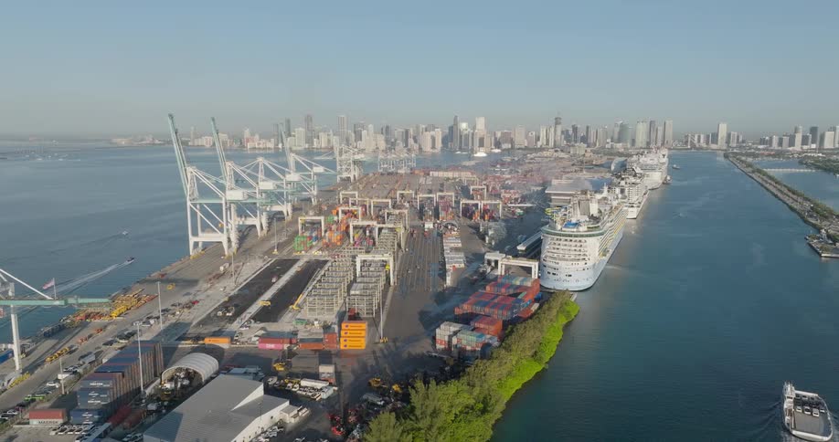 An aerial view shows the busy Port of Miami with its cargo cranes, shipping containers, and docked cruise ships with the city skyline in the background