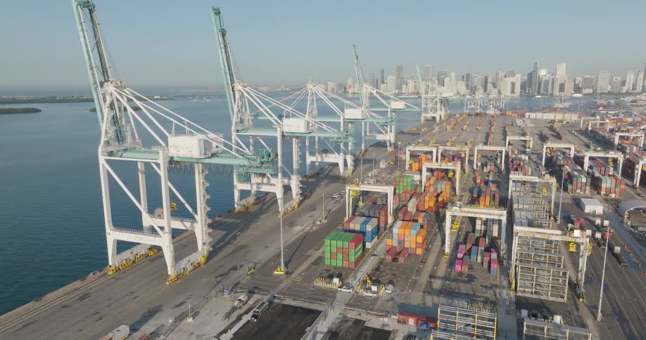 A sweeping aerial pullback shot reveals the massive cranes and colorful shipping containers at the Port of Miami, with the city skyline in the background
