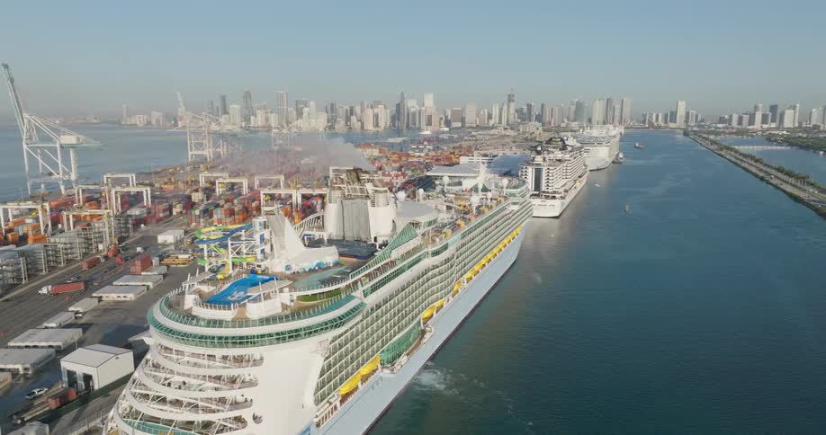 A stunning aerial view shows a massive Royal Caribbean cruise ship docked at the bustling Port of Miami, with the city skyline visible in the background