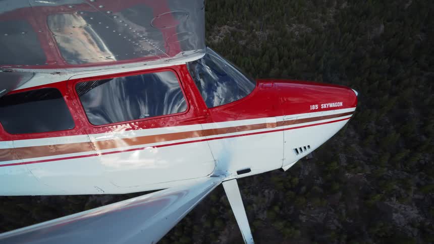 Aerial view from the wing of a red and white 185 Skywagon plane as it flies over a forested mountain range