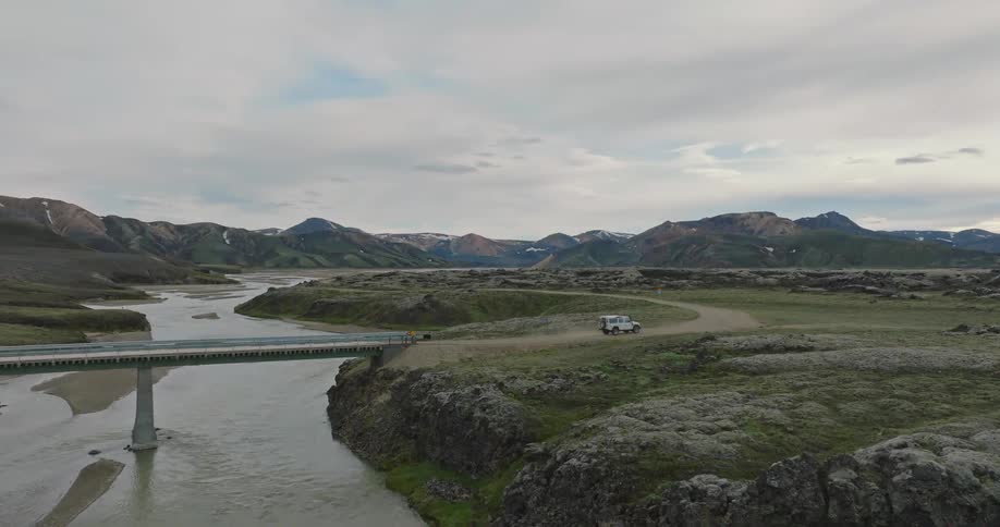 An aerial view shows a white 4x4 vehicle crossing a bridge over a river and driving on a dirt road through the remote and scenic Icelandic highlands