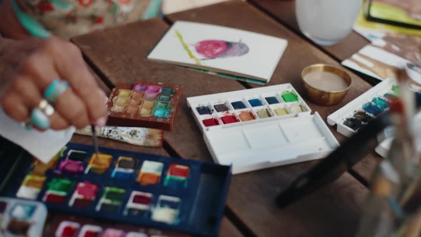 An artist's hands delicately apply watercolor paint to a small canvas, bringing a colorful bird to life