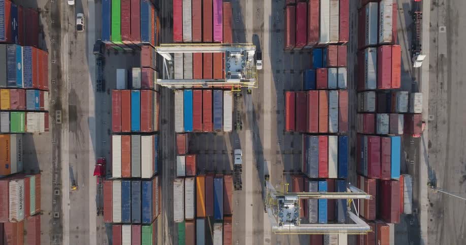 Top-down aerial view slowly ascends over a bustling shipping container port, showing organized rows of colorful containers, moving cranes, and trucks