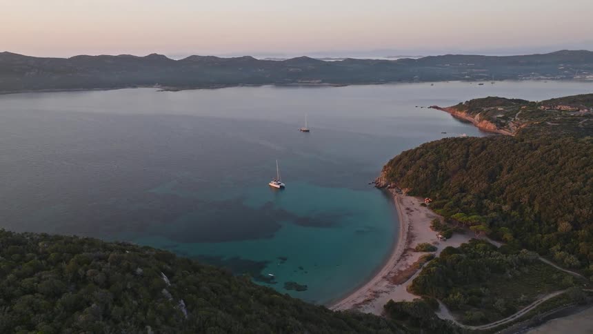 A picturesque aerial shot shows sailboats resting in a calm, turquoise bay next to a secluded sandy beach during a beautiful sunrise