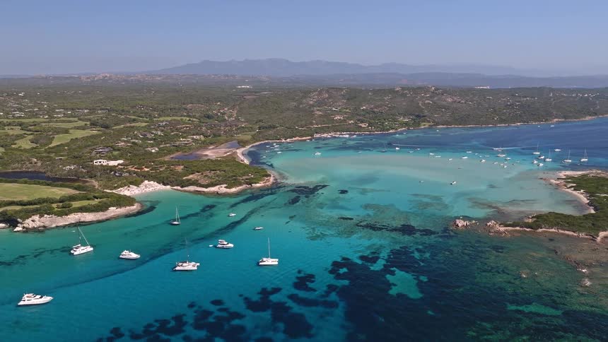 An ascending aerial view showcases luxury yachts and sailboats peacefully anchored in the stunningly clear, turquoise waters of a picturesque coastal bay