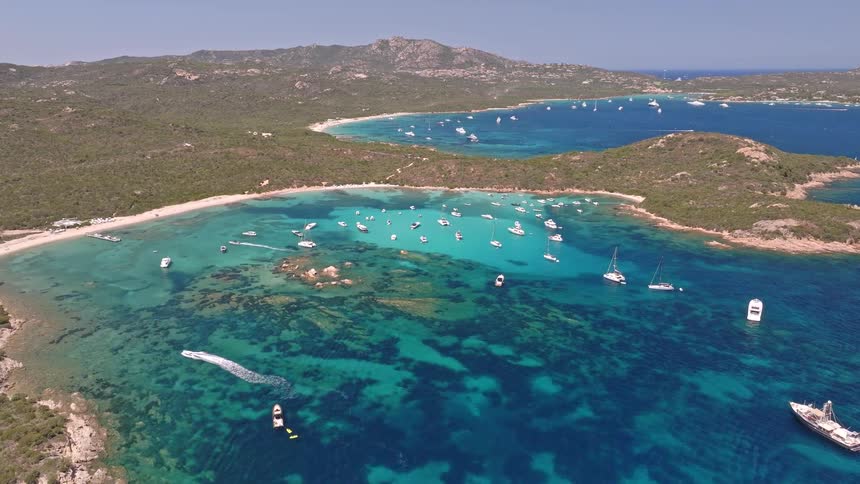 A static aerial view of a picturesque Mediterranean cove where numerous yachts and sailboats are anchored in the stunningly clear turquoise water