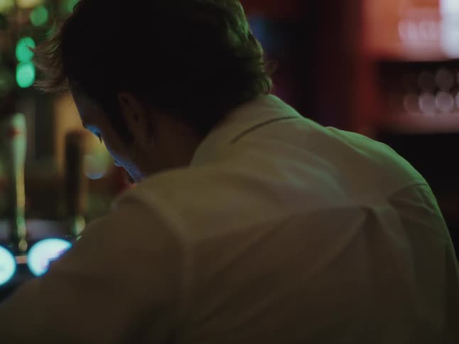 An over-the-shoulder view shows a man in a white shirt sitting at a dimly lit bar