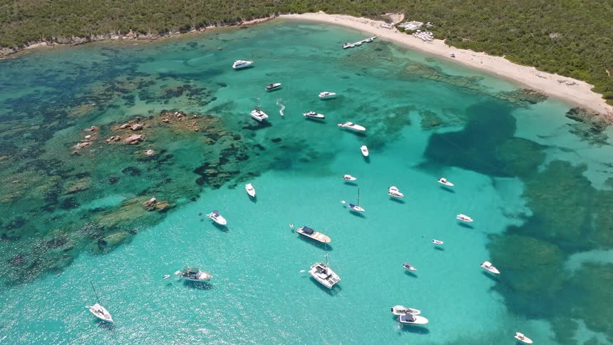 Stunning aerial flight over dozens of luxury yachts and boats floating in a pristine turquoise bay surrounded by a lush green island coastline