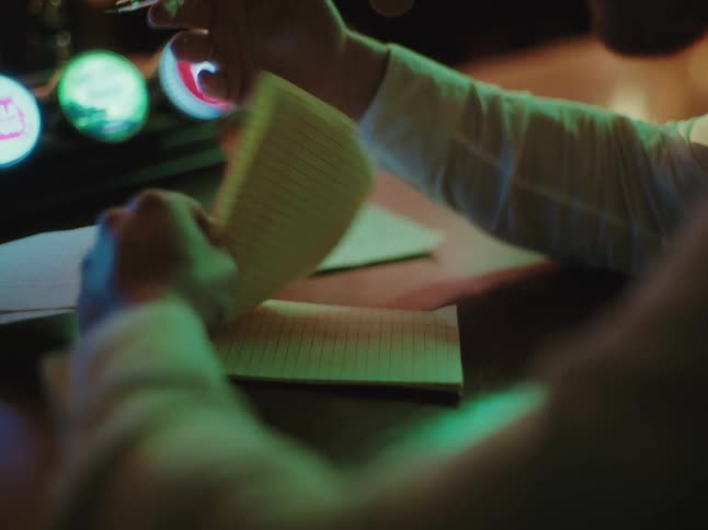 A person's hands flip through the pages of a notebook and write with a pen while sitting at a dimly lit bar counter