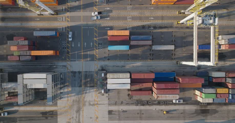 Top-down aerial view of a bustling shipping container terminal with gantry cranes moving along tracks and vehicles transporting cargo