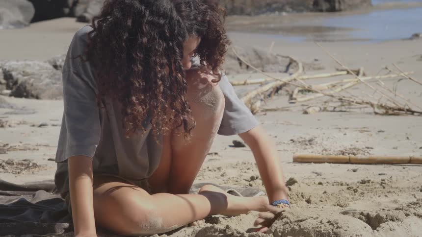 A thoughtful young woman with curly hair plays with the sand on a sunny beach