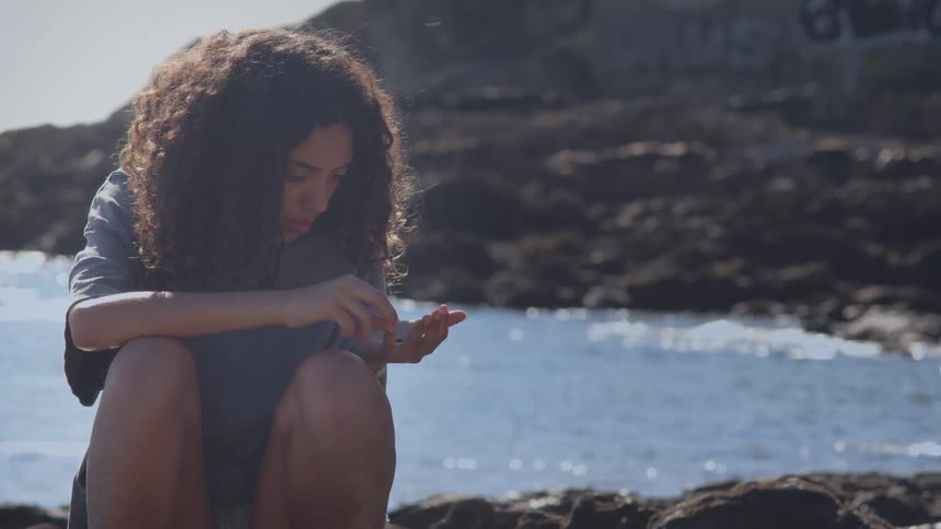 A young person with curly hair sits on a rocky shore, thoughtfully examining a small object they found by the sparkling water