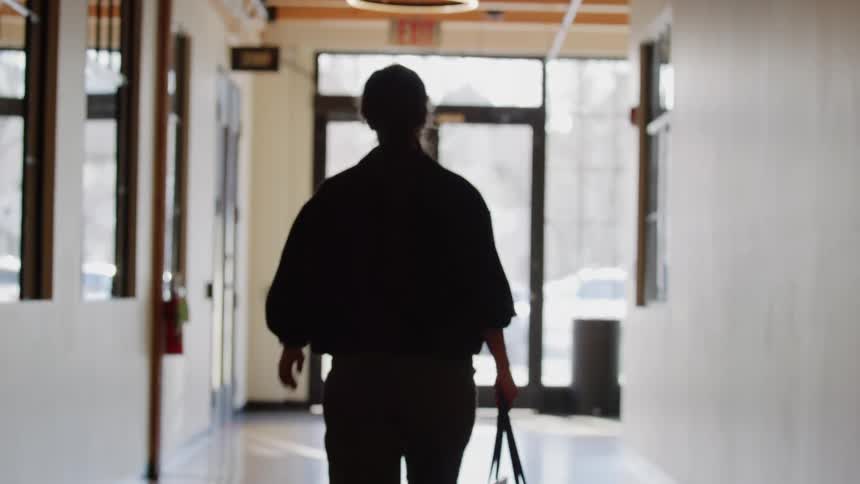 A camera pans from a modern circular light fixture to follow a silhouetted person walking down a long hallway