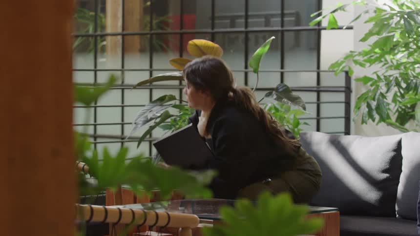 A woman finishes her work, closes her laptop, and leaves a modern lounge filled with lush green plants