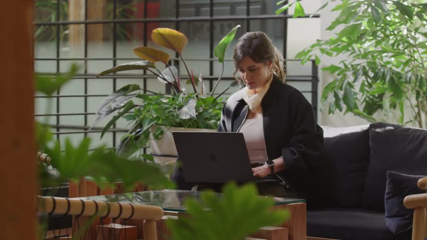 A woman works remotely on her laptop while sitting on a comfortable sofa in a bright, modern office space filled with lush green plants
