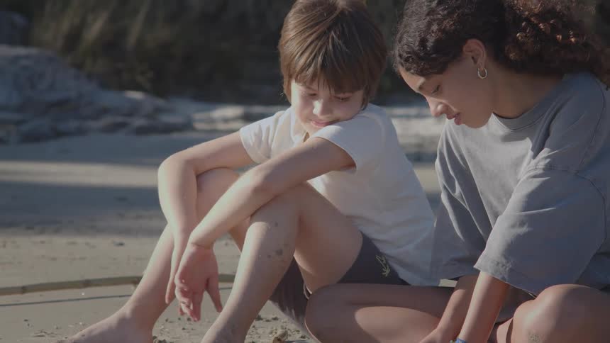 A young boy and his older sister sit on a sandy beach, happily playing together in the warm sunlight