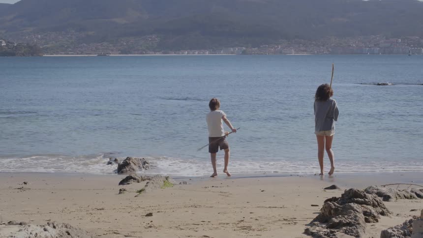 A young boy and girl stand at the edge of the ocean on a sandy beach playing with long sticks in slow motion