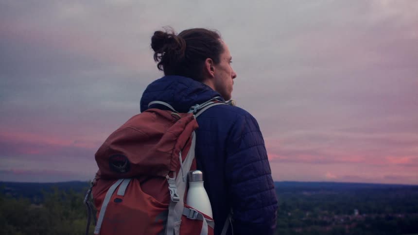A hiker with a backpack stands on a hilltop, gazing out at the vast landscape under a beautiful pink and purple sunset sky