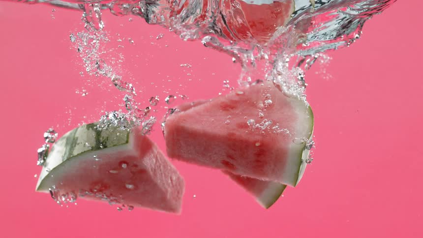 Watermelon slices plunge into water in super slow motion, splashing and creating bubbles against a vibrant pink background