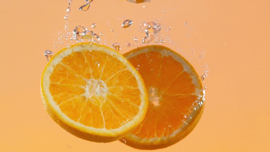 Two fresh orange slices slowly submerge in clear water against an orange background in slow motion