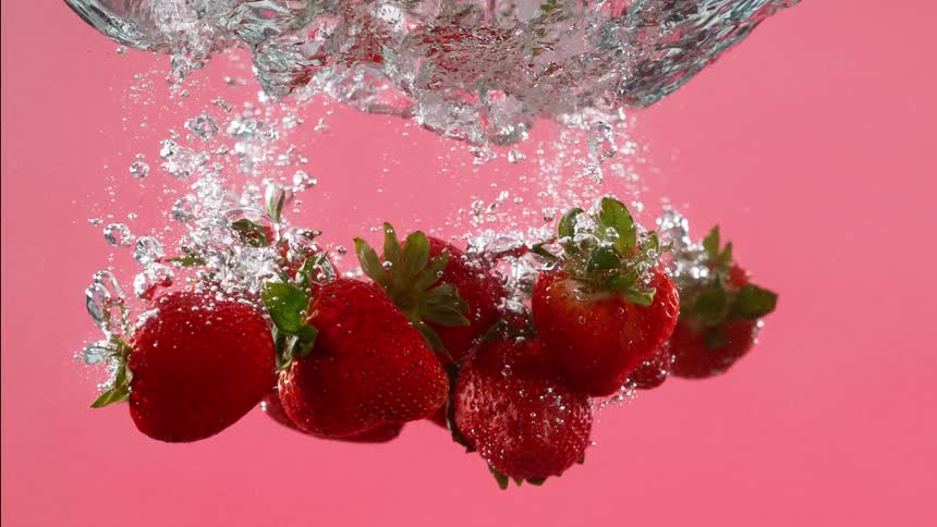A handful of fresh strawberries drop into water in super slow-motion against a solid pink background