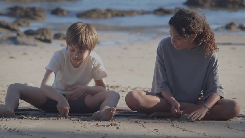 Two happy children sit cross-legged on a sandy beach, drawing pictures in the sand with their fingers by the ocean