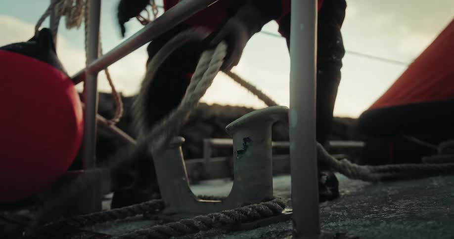 A sailor in a red waterproof suit unties a thick mooring rope from a cleat on the frosty deck of a boat in a harbor