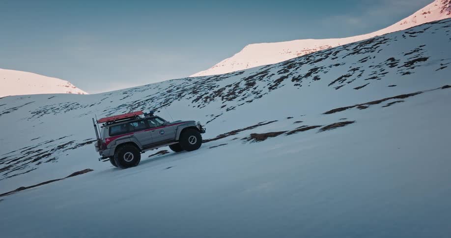 A rugged 4x4 super jeep slowly ascends a steep, snow-covered mountainside in a vast and remote winter landscape