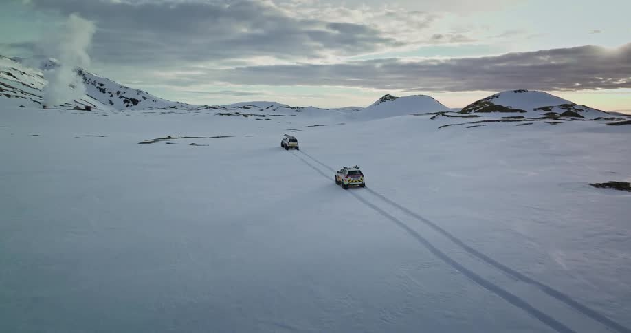 An aerial shot follows two expedition vehicles as they drive through a vast, snow-covered landscape with mountains and geothermal steam in the background