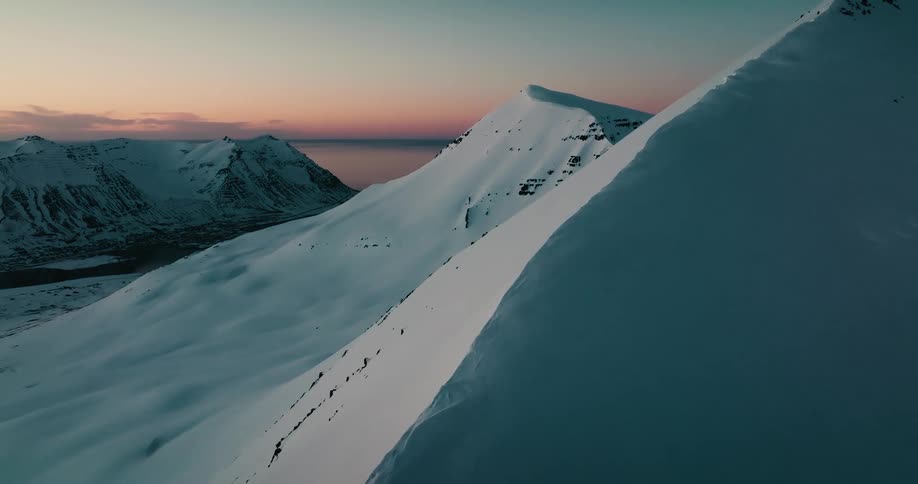 An aerial reveal shot pulls back from a snowy mountain ridge to show a vast, scenic winter landscape at sunrise