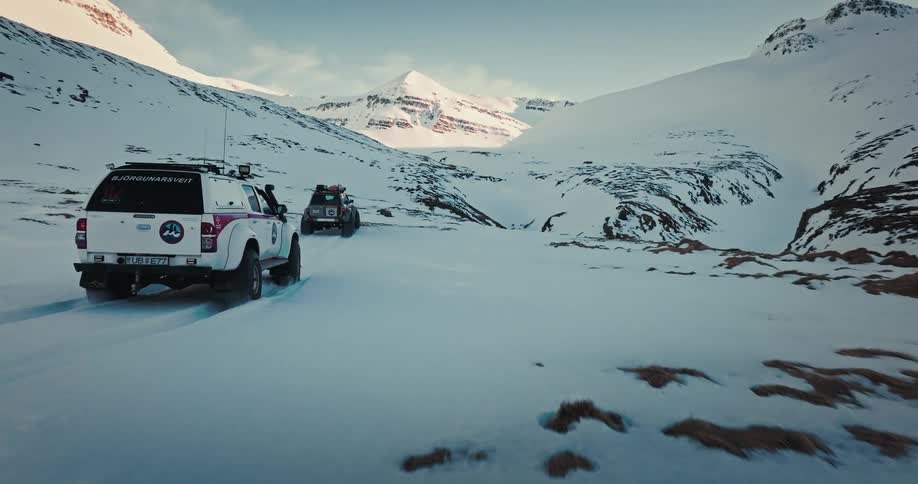 Cinematic drone shot follows two rugged expedition vehicles as they drive through a vast, snow-covered mountain valley