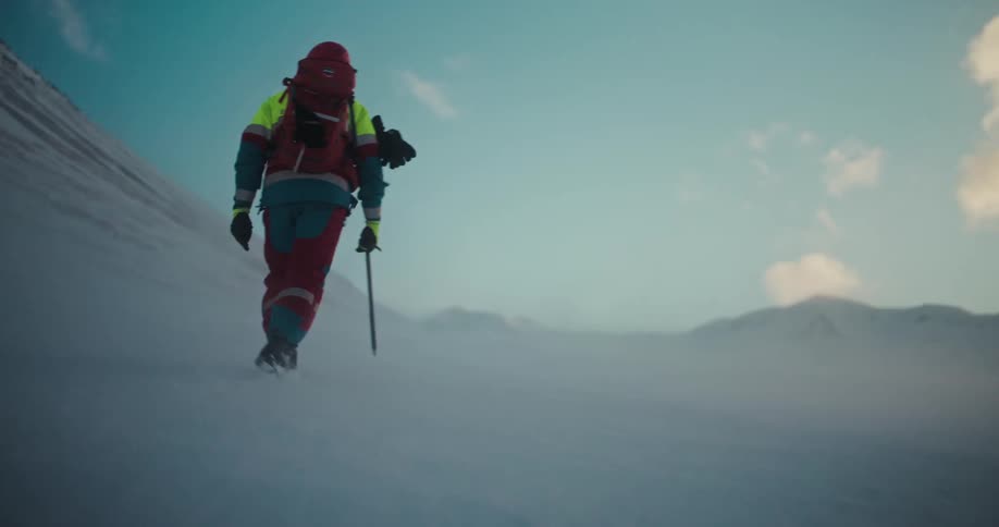 A dramatic slow-motion shot follows two fully-equipped mountaineers as they ascend a vast, windy, and snow-covered mountain