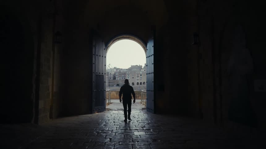 A silhouetted man walks in slow motion through a dark, ancient stone gateway and out into the light revealing a city view