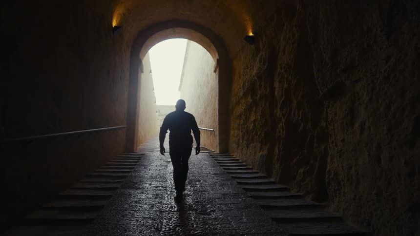 A man's silhouette walks in slow motion down a dark, wet, stone tunnel toward a bright light at the end